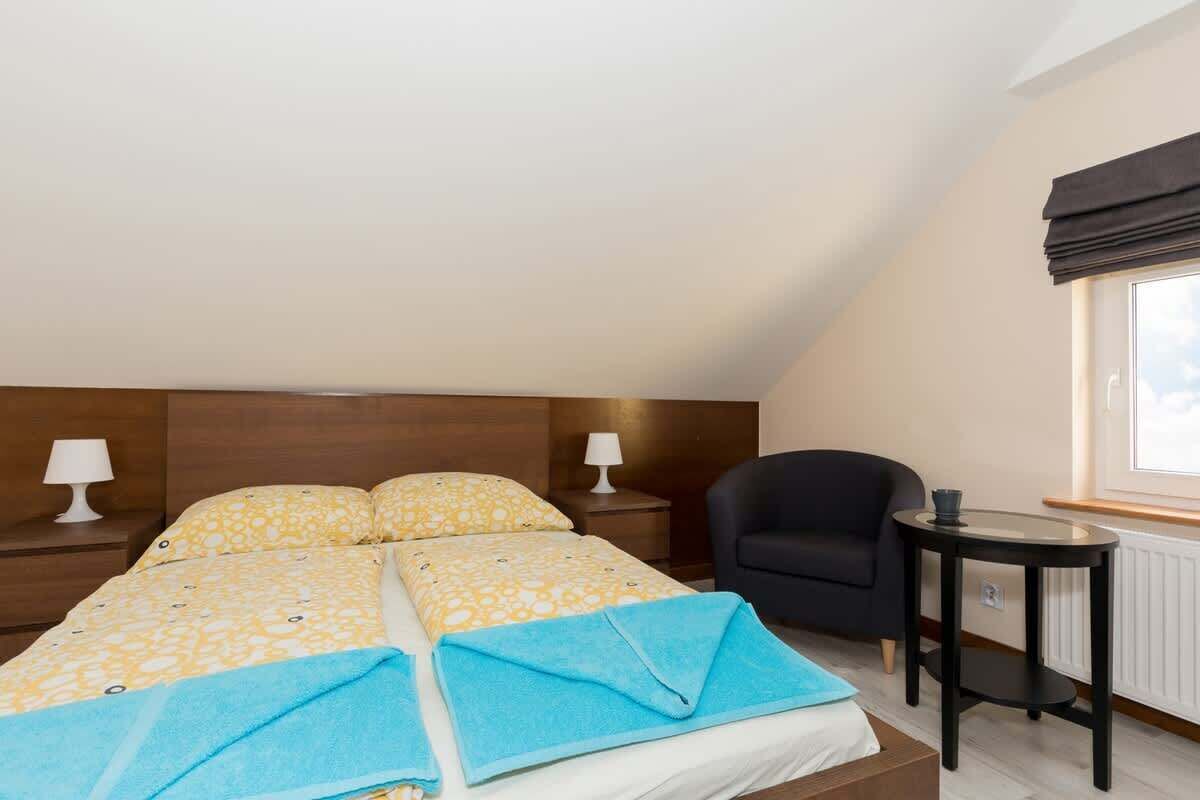 A bedroom with a bed, two bedside tables with lamps, a black armchair, a small round table, and a window with a view.