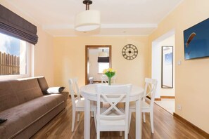 A room with a beige sofa, a white dining table with four chairs, a clock on the wall, and a window with blinds.