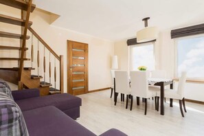A modern living room with a staircase, a dining area with white chairs, and a large window with a view.