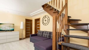 Una sala de estar con un sofá morado, una escalera de madera, un reloj de pared y un televisor sobre un mueble.