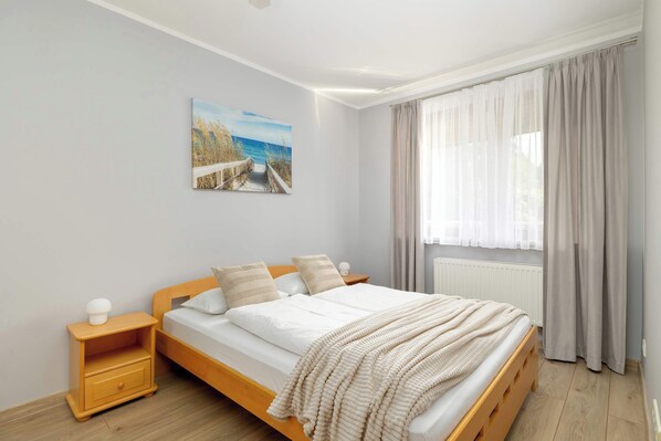 A bedroom with a wooden bed, a nightstand, a window with curtains, and a beach scene painting on the wall.