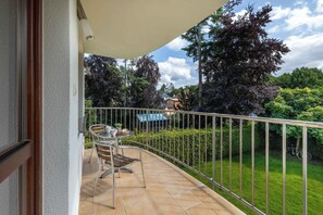 Ein Balkon mit Metallgeländer, einem Tisch mit Stühlen und Blick auf eine grüne Rasenfläche und Bäume.