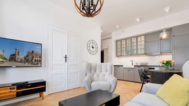 Una sala moderna con una televisión de pantalla plana, un sofá, una mesa de centro y una silla. Hay una zona de cocina con armarios grises y una mesa de comedor.