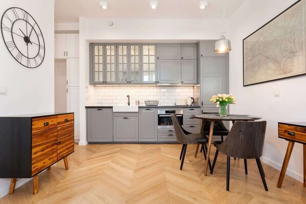 Une cuisine moderne avec des meubles gris, un parquet en bois disposé en motif chevron, une horloge noire et un coin repas comprenant deux chaises et une table.