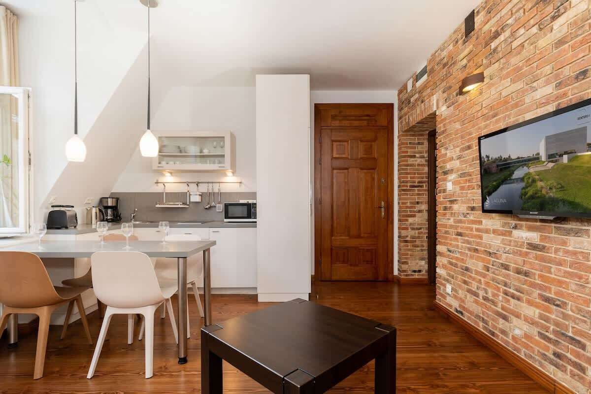 A modern kitchen with white cabinets, a dining area with a table and chairs, and a brick wall with a mounted TV.