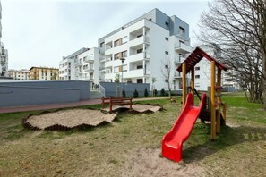 A playground with a red slide and wooden play structures, surrounded by a grassy area and a building with multiple balconies in the background.