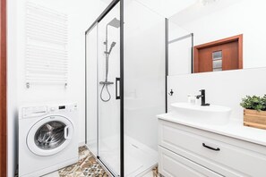 A modern bathroom with a glass-enclosed shower, a built-in washing machine, and a white sink with a wooden countertop.