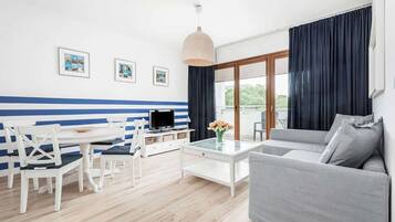 Een moderne woonkamer met een witte bank, een glazen salontafel en een eethoek met een witte tafel en stoelen. Er hangen ingelijste foto’s aan de muur en er is een groot raam met gordijnen.