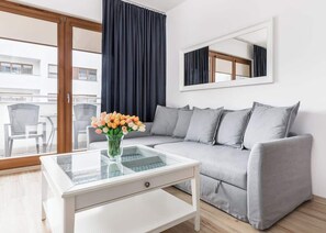 Un salon moderne avec un canapé gris, une table basse en verre ornée d’un vase de fleurs, un grand miroir au mur et une vue sur le balcon extérieur.