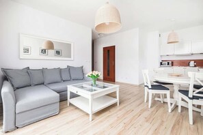 Un salon moderne avec un canapé gris, une table basse blanche ornée d’un vase de fleurs et un coin repas avec des chaises blanches et une table en bois.