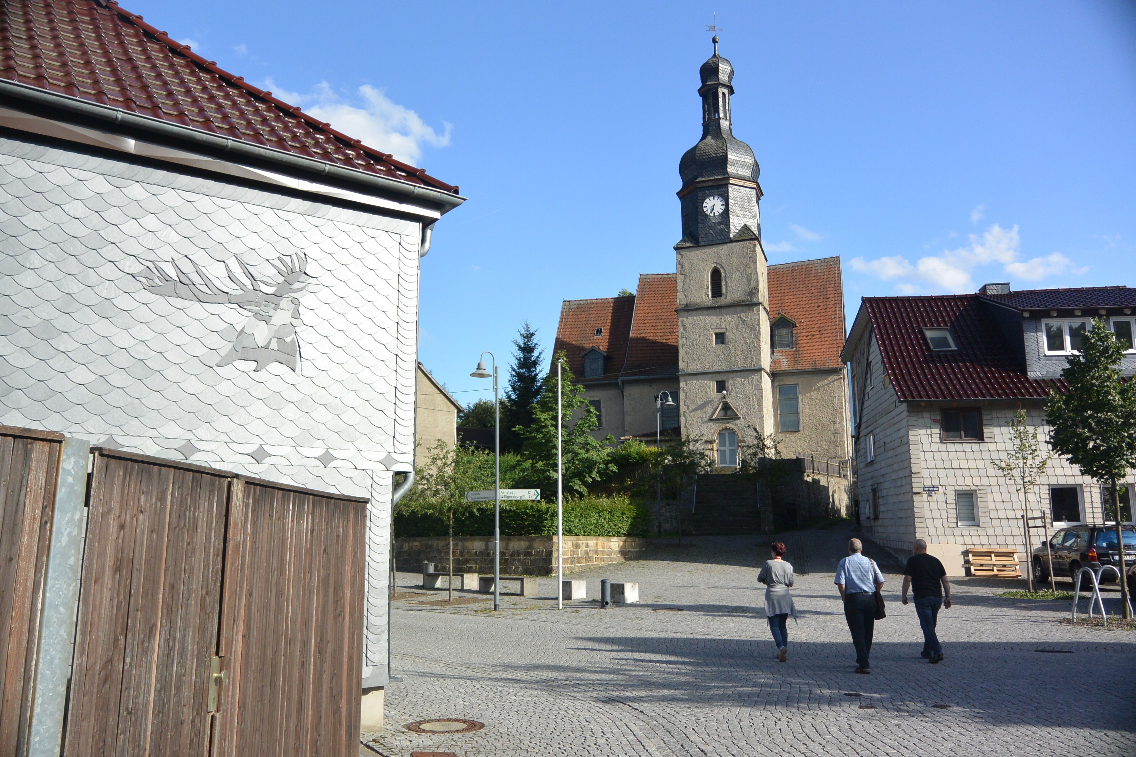 A cobblestone square with a church tower, a wooden building with a dragon mural, and a clear blue sky.