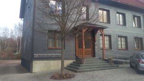 Un edificio de dos pisos con fachada gris y una puerta de madera. Hay un árbol frente al edificio, y un coche aparcado en la entrada.