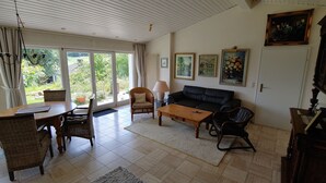 A living room with a sofa, coffee table, chairs, a lamp, and a dining table. There are large windows and a view of greenery outside. - Beautiful detached holiday home with a sunny location and panoramic views (Lissendorf)