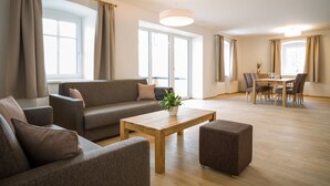 Een moderne woonkamer met een bank, een voetenbankje en een salontafel, een eethoek met stoelen en een tafel, en grote ramen met gordijnen.