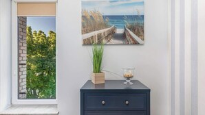 A room with a view of a beach through a window, a dark blue dresser, a painting of a beach scene, and a potted plant.