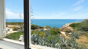 Une vue sur la côte, depuis une fenêtre donnant sur une plage de sable, un ciel bleu dégagé et un paysage rocheux parsemé de végétation.