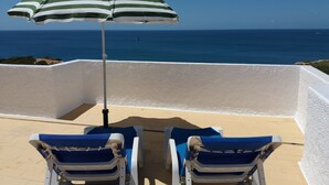 Deux fauteuils de jardin avec un parasol à rayures, une vue sur la mer et un mur blanc.