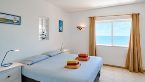 Une chambre à coucher avec un lit, une table de chevet, une fenêtre avec des rideaux et une vue sur la mer.