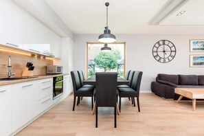 A modern kitchen with white cabinets, a dining area with a wooden table and black chairs, and a living area with a dark sofa and a wooden coffee table.