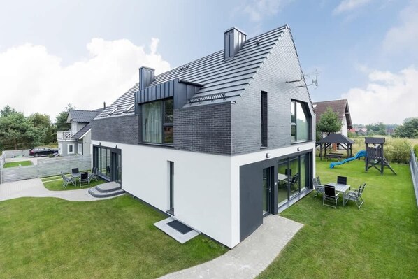 Une maison moderne à deux étages, dotée d’un toit en pente, de grandes fenêtres et d’un espace de jeux bien visible, avec un toboggan.