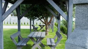 A covered picnic area with two wooden benches and a table, surrounded by grass and trees.