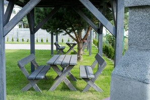 A covered picnic area with two wooden benches and a table, surrounded by grass and trees.