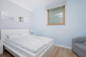 A bedroom with a bed, a nightstand, a window with a wooden frame, and a grey sofa.