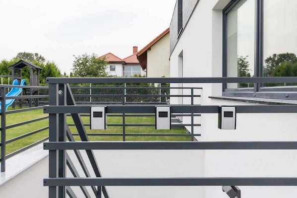 A modern building with a balcony, a fenced play area with a slide, and a house in the background.