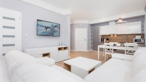 A modern living room with white sofas, a dining area, and a kitchen with white cabinets and a wooden countertop.