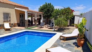 Una piscina con sillones de descanso y una pérgola, rodeada por un jardín y un edificio con techo de tejas.