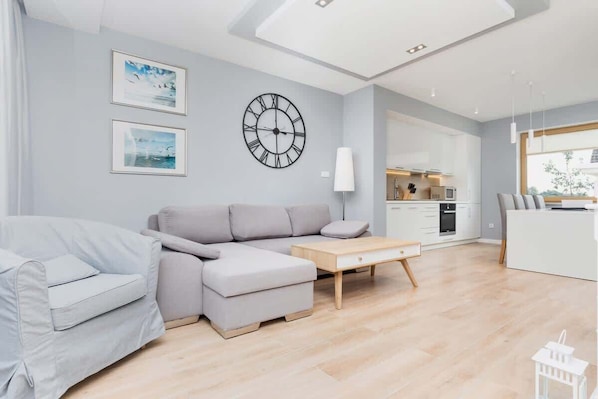 A modern living room with a grey sectional sofa, a wooden coffee table, and a large wall clock. There are framed pictures on the wall, a white lamp, and a view of the kitchen in the background.