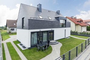 Une maison moderne à deux étages, dotée d’un toit en pente, de grandes fenêtres et d’une terrasse aménagée avec du mobilier d’extérieur.