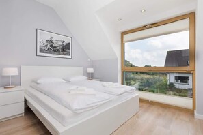 A modern bedroom with a large bed, two bedside tables, a framed picture on the wall, and a large window offering a view of the outside.