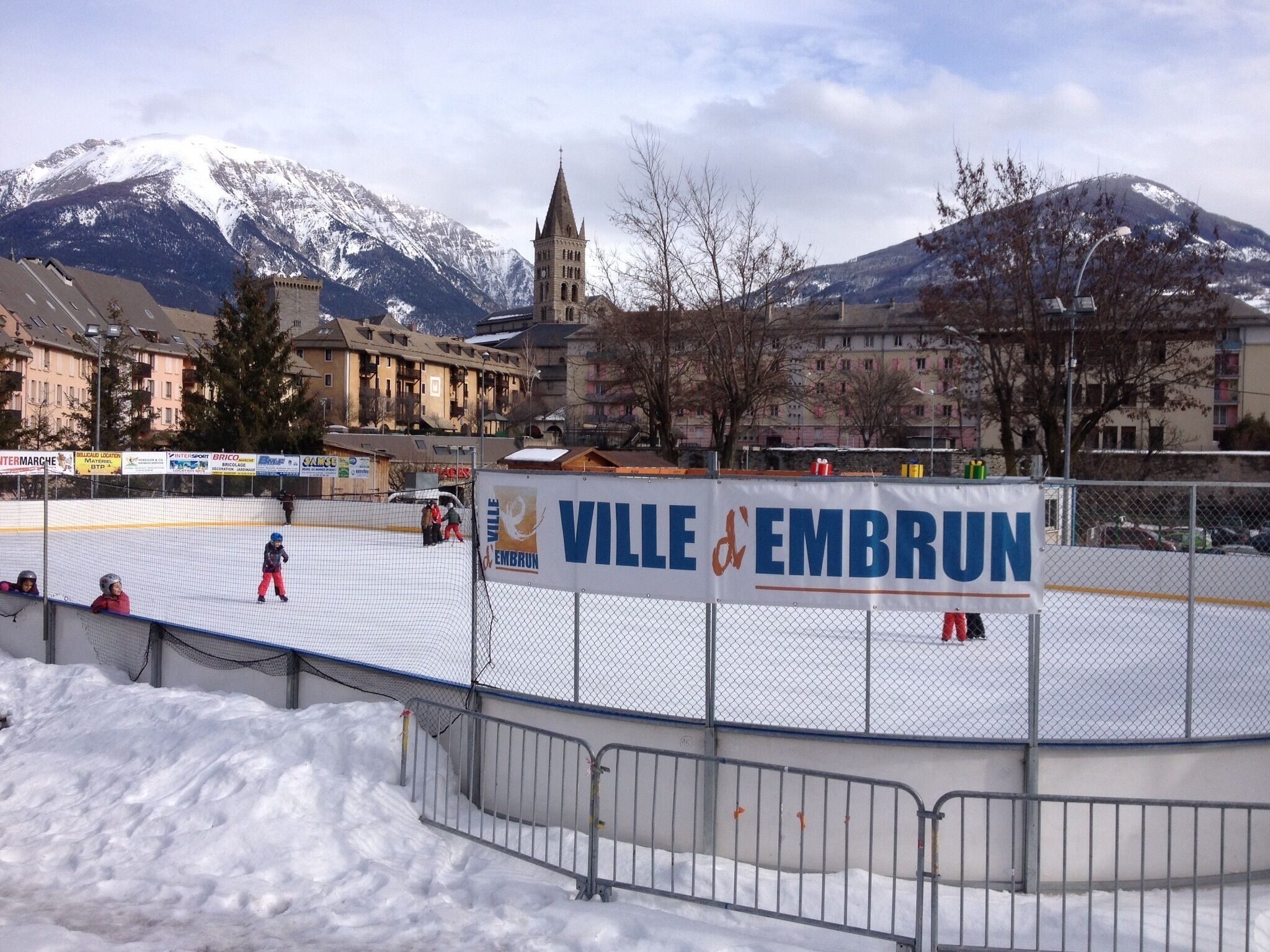 Eine Eisbahn im Freien mit einer Aufschrift 'VILLE & EMBRUN' und einem Bergpanorama im Hintergrund.