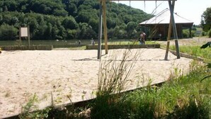 A sandy beach with a wooden swing set and a covered area, surrounded by greenery and a forested hill. - Cottage of the Barry fountain in Aveyron (Le Vibal)