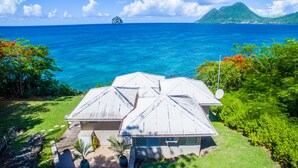 Una casa con techo blanco y antena parabólica, rodeada de vegetación exuberante y con vistas al mar azul cristalino.