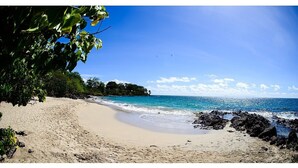 Una playa con agua azul cristalina, orilla arenosa y algunas rocas cerca del agua.