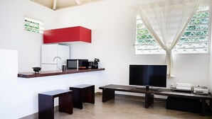 Una cocina moderna con un refrigerador blanco, microondas y cafetera sobre una encimera de madera. Hay un televisor de pantalla plana grande sobre un soporte, un banco y una ventana con cortinas blancas.