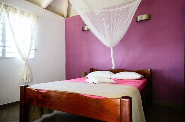 Ein Schlafzimmer mit einem hölzernen Bett, weißer Bettwäsche, einem weißen Moskitonetz und violetten Wänden.