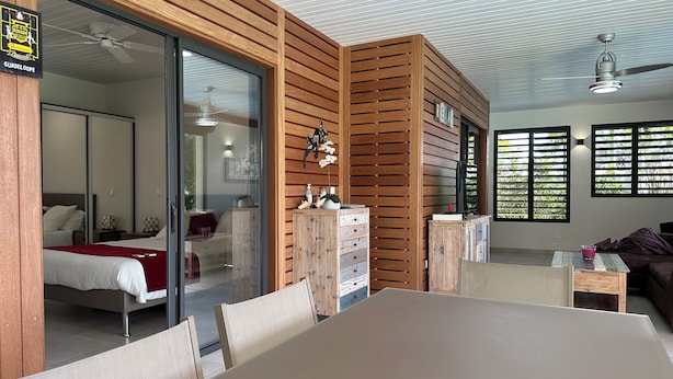 Une chambre d’hôtel moderne avec un mur d’accent en bois, une porte-fenêtre coulissante donnant sur un balcon, un lit avec une couverture rouge, une petite table avec une boisson rose et un ventilateur de plafond.