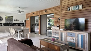Un salon moderne avec un mur d’accent en bois, un téléviseur et un coin repas, la cuisine étant visible au fond.