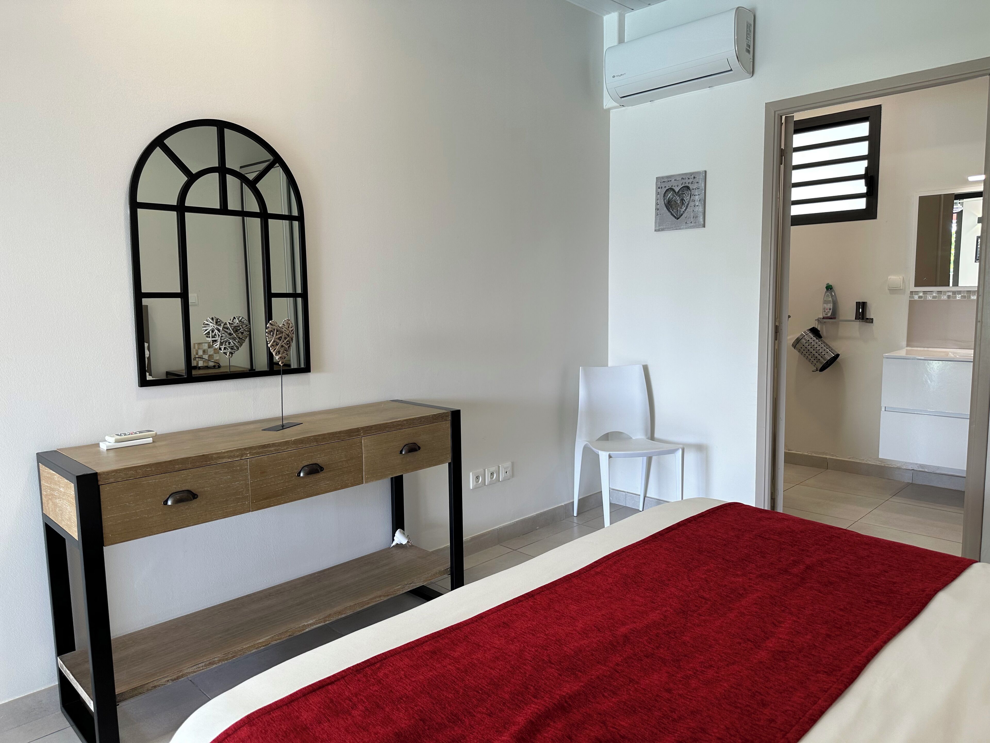 Un dormitorio con cama, una consola de madera con cajones, una silla blanca, un espejo y una manta roja.