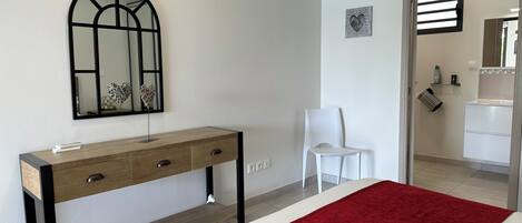 Un dormitorio con cama, una consola de madera con cajones, una silla blanca, un espejo y una manta roja.