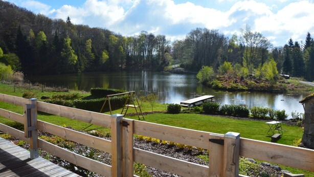 Une terrasse en bois donnant sur un lac, avec un banc et une balançoire, le tout entouré d’une végétation luxuriante et d’arbres.