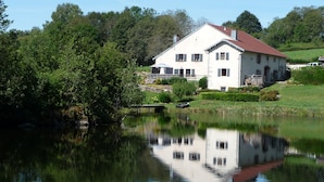 Une grande maison blanche coiffée d’un toit rouge, entourée d’une végétation luxuriante et bordée d’un étang paisible dans lequel se reflète son image.