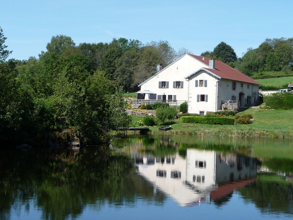 A large white house with a red roof, surrounded by lush greenery and a calm pond reflecting its image.