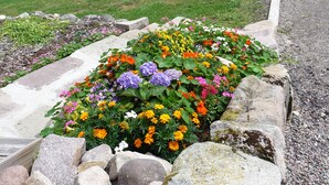 Un bâtiment rustique en pierre, doté d’une porte en bois, entouré d’un parterre fleuri avec différentes fleurs aux couleurs vives, d’un chemin de gravier et, au fond, d’un étang.