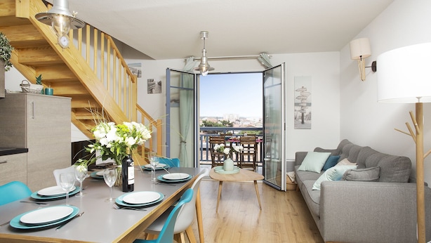 A modern kitchen with a wooden staircase, a dining area with blue chairs, and a grey sofa.