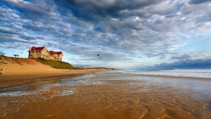 A beach with a large building on a hill, waves approaching the shore, and a partly cloudy sky.