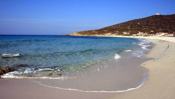Een strand met helderblauw water, een zandstrand en een heuvel met schaarse begroeiing.